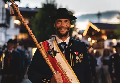 A man in traditional attire is holding a long staff in his hand. In the background, people in traditional costumes and festive lighting can be seen.