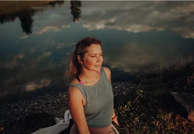 A woman is sitting by the shore of a lake, thoughtfully gazing into the water. The surroundings are reflected in the calm water, creating a peaceful atmosphere.