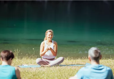 A group of people is doing yoga by the shore of a calm lake. A woman sits smiling in the foreground and leads the exercise.