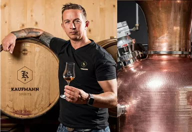 A professional stands next to wine storage barrels and holds a glass in hand. In the background, copper distillation equipment can be seen.