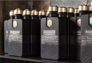 A series of black bottles with golden caps is standing on a shelf. The bottles are labeled "KAUFMANN" and appear modern and elegant.