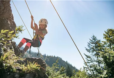 A child is climbing a rock wall and using a rope for safety. The surroundings are filled with trees and mountains, and the sky is clear and sunny.