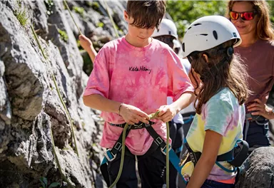 Children are preparing for climbing while wearing helmets. They are standing at a rock wall and are equipped with safety gear.