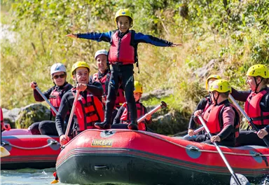 Eine Gruppe von Menschen in einem Schlauchboot auf einem klaren Fluss. Ein Kind steht mit ausgestreckten Armen an der Vorderseite des Bootes.