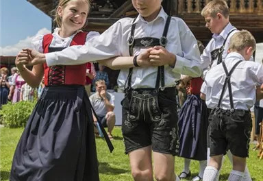 A joyful dance of children in traditional Bavarian costume. In the background stands a wooden house and the sun is shining.