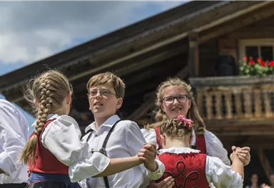Kinder in traditioneller Tracht tanzen zusammen vor einem rustikalen Holzhaus. Der Himmel ist blau und die Stimmung ist fröhlich.