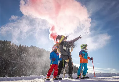 Ein fröhlicher Wintersporttag mit Kindern, die auf einem verschneiten Hang stehen. Eine Person im Kostüm zeigt mit einem farbigen Rauchgranat in den Himmel.