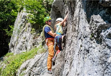 A climber helps a child climb a rock wall. The sun is shining, and the surroundings are green and natural.
