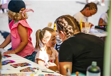 Ein Kind bekommt ein Gesichtsmalerei von einer Betreuerin. Im Hintergrund sind weitere Kinder beim Malen zu sehen.