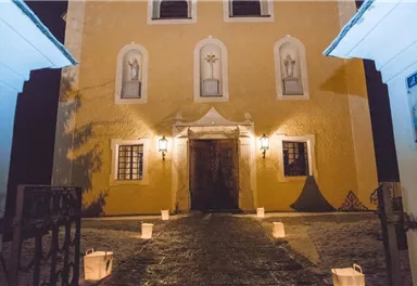A historic church with a yellow facade and illuminated windows. The path to the entrance door is lined with lanterns.