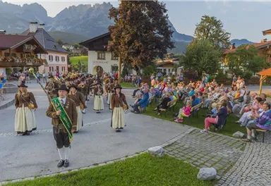 Ein traditionselles Fest mit einer Blaskapelle und tanzenden Menschen in Trachten. Im Hintergrund sitzt ein begeistertes Publikum auf Stühlen.