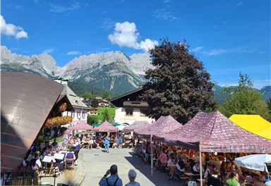 Ein lebhaftes Fest mit vielen Zelten und Besuchern in einer malerischen Berglandschaft. Der Himmel ist klar und die Umgebung ist von Natur umgeben.