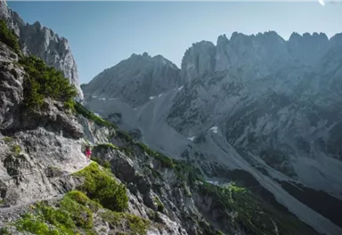 Eine beeindruckende Gebirgslandschaft mit hohen Felsen und schneebedeckten Gipfeln. Ein Wanderer folgt einem schmalen Pfad zwischen den Bergen.