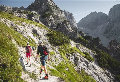 Zwei Wanderer gehen einen steilen Pfad in den Bergen. Die Landschaft ist beeindruckend mit hohen Felsen und grünen Wiesen.