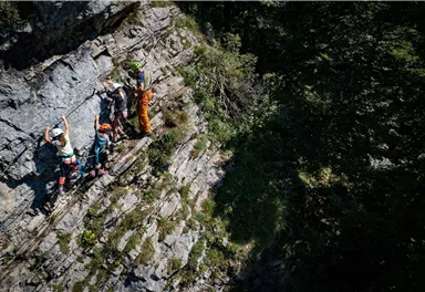 A group of climbers is moving along a steep rock face. The surroundings are green and forested, which emphasizes the natural beauty of the scene.