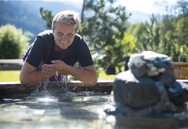 Eine Frau lehnt sich über einen kleinen Wasserfall und lächelt in die Kamera. Im Hintergrund sind grüne Bäume und eine hügelige Landschaft zu sehen.