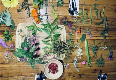 A wooden board with various herbs and edible flowers lies on a table. Around the board are several place settings with cutlery arranged.