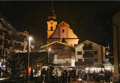 A festive marketplace at night with snowfall. In the background, a church and illuminated buildings can be seen.