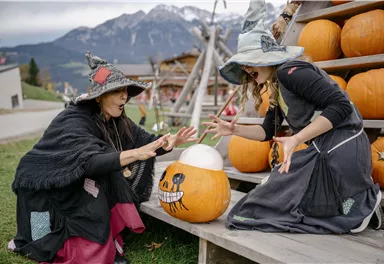 Zwei Frauen in Hexenkostümen stehen vor einem Holzregal mit Kürbissen. Sie scheinen einen Kürbis mit einem Gesicht zu betrachten und wirken dabei begeistert.