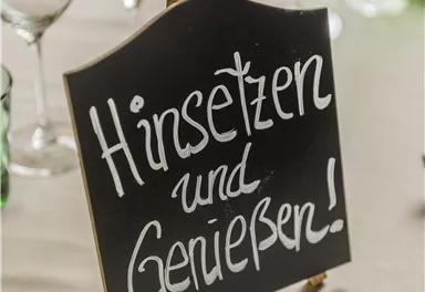 A sign with the inscription "Sit down and enjoy!" stands on a table. In the background, glasses and colorful decorative elements can be seen.