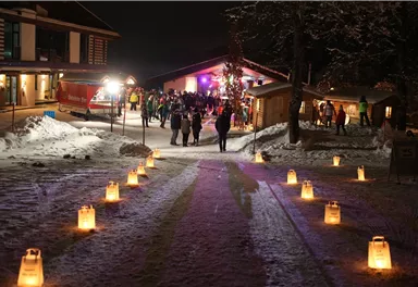 A festively illuminated winter landscape with snow-covered paths. Groups of people gather around a cozy cabin under the night sky.
