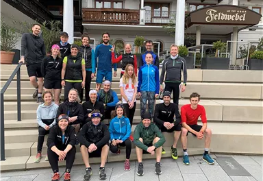 Eine Gruppe von Menschen steht auf Treppenstufen vor einem Hotel. Alle sind sportlich gekleidet und posieren freundlich für das Foto.