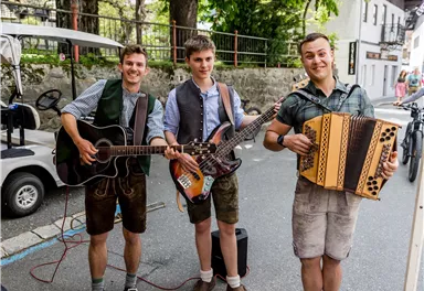 Three men in traditional clothing are playing musical instruments on a street. They have an accordion, a guitar, and an electric bass.