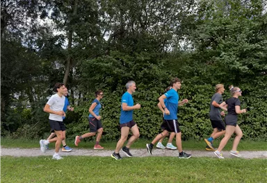 Eine Gruppe von Menschen joggt auf einem schmalen Weg. Umgeben von viel Grün und Bäumen bewegen sie sich in einem aktiven Tempo.