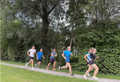 Eine Gruppe von sechs Personen joggt auf einem Kiesweg entlang dichter Bäume. Alle Teilnehmer tragen Sportkleidung und wirken aktiv und energiegeladen.