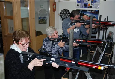 A group of people is aiming with sporting rifles in a training room. The atmosphere is focused and athletic.