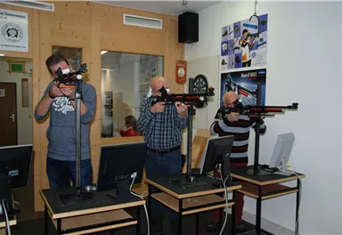 Three men stand at shooting ranges, aiming with air rifles. In the background, computers and a bulletin board are visible.