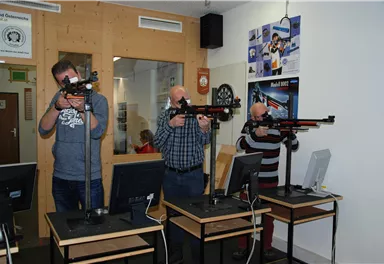 Three men are standing at shooting ranges with rifles. They are focused on aiming and shooting, while computers are in front of them.