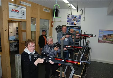 A group of five people stands in a room and conducts training sessions with sports rifles. Offices and posters are visible in the background.