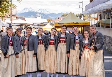 A group of girls and women in traditional costumes stand smiling together. In the background, mountains and a festival tent can be seen.