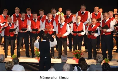 Ein Männerchor steht auf der Bühne und singt. Sie tragen rote Westen und schwarze Hosen, während das Publikum aufmerksam zuhört.