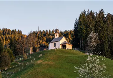 A small church surrounded by trees and mountains. The sky is clear and blue, creating a peaceful atmosphere.