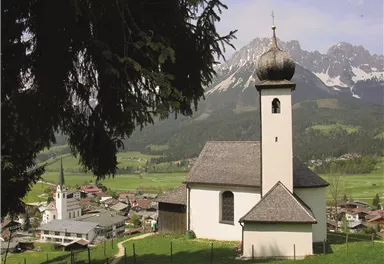 A picturesque church with a distinctive dome stands in a green landscape. In the background, majestic mountains can be seen framing the idyllic scene.