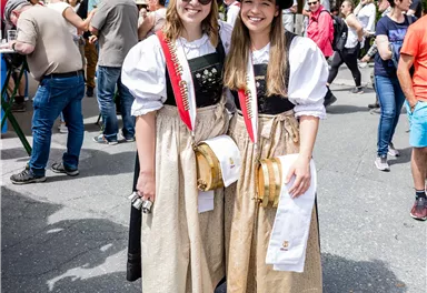 Two girls in traditional costumes with black hats and golden skirts. They are smiling happily in front of a crowd at a festival.