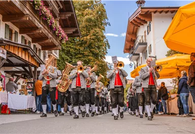 Eine Musikkapelle in traditionellen Trachten marschiert durch eine festliche Straße. Im Hintergrund sind Stände und bunte Sonnenschirme zu sehen.