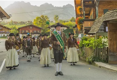 Eine festliche Parade mit einer Gruppe von Menschen in traditioneller Tracht. Im Hintergrund sind malerische Berge und Holzhäuser zu sehen.