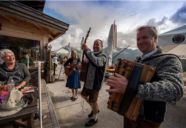 Eine fröhliche Musikkapelle unterhalb eines Bergs spielt traditionelle Lieder. Gäste genießen die Atmosphäre in einem gemütlichen Außenbereich.