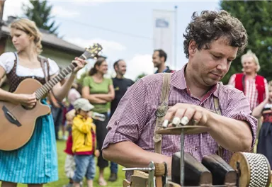 A festive atmosphere with people enjoying music. A man plays a traditional musical instrument while a woman sings with a guitar.