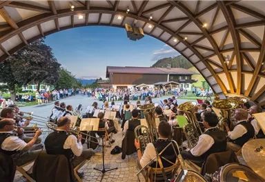 Eine Blaskapelle spielt vor einer großen Menschenmenge unter einem gewölbten Dach. Im Hintergrund sind Berge und ein klarer Himmel sichtbar.