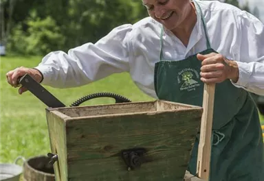 A man is working on an old juice press outdoors. In the background, there are trees and a green meadow.