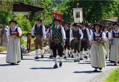 Eine traditionelle Trachtenparade mit Menschen in festlicher Kleidung. Sie gehen fröhlich eine Straße entlang, umgeben von Bäumen und ländlicher Umgebung.