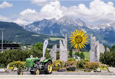 Ein grüner Traktor steht vor einem sonnigen, blühenden Blumenbeet. Im Hintergrund sind majestätische Berge und ein strahlend blauer Himmel zu sehen.