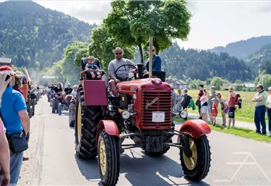 Eine Traktorrallye mit vielen Teilnehmenden auf Traktoren in einer ländlichen Umgebung. Die Menschen stehen am Straßenrand und beobachten das Event.