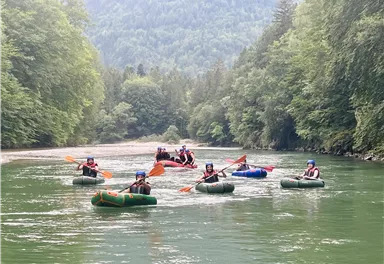 Eine Gruppe von Personen fährt mit Schlauchbooten auf einem ruhigen Fluss. Umgeben von grünen Bäumen und Bergen bietet die Szenerie eine entspannte Atmosphäre.