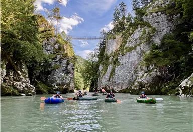 Eine Gruppe von Menschen schwimmt in einem Fluss zwischen hohen Felsen. Im Hintergrund ist eine Hängebrücke sichtbar, umgeben von grünem Wald.