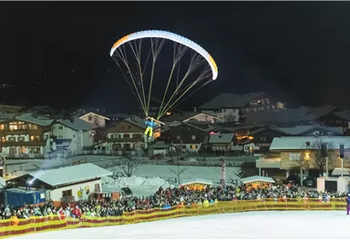 A paraglider glides over a crowd at night in a snow-covered village. In the background, festively lit houses can be seen.